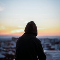 A solitary figure in a hoodie gazes at a vibrant sunset from a rooftop, creating a peaceful silhouette.